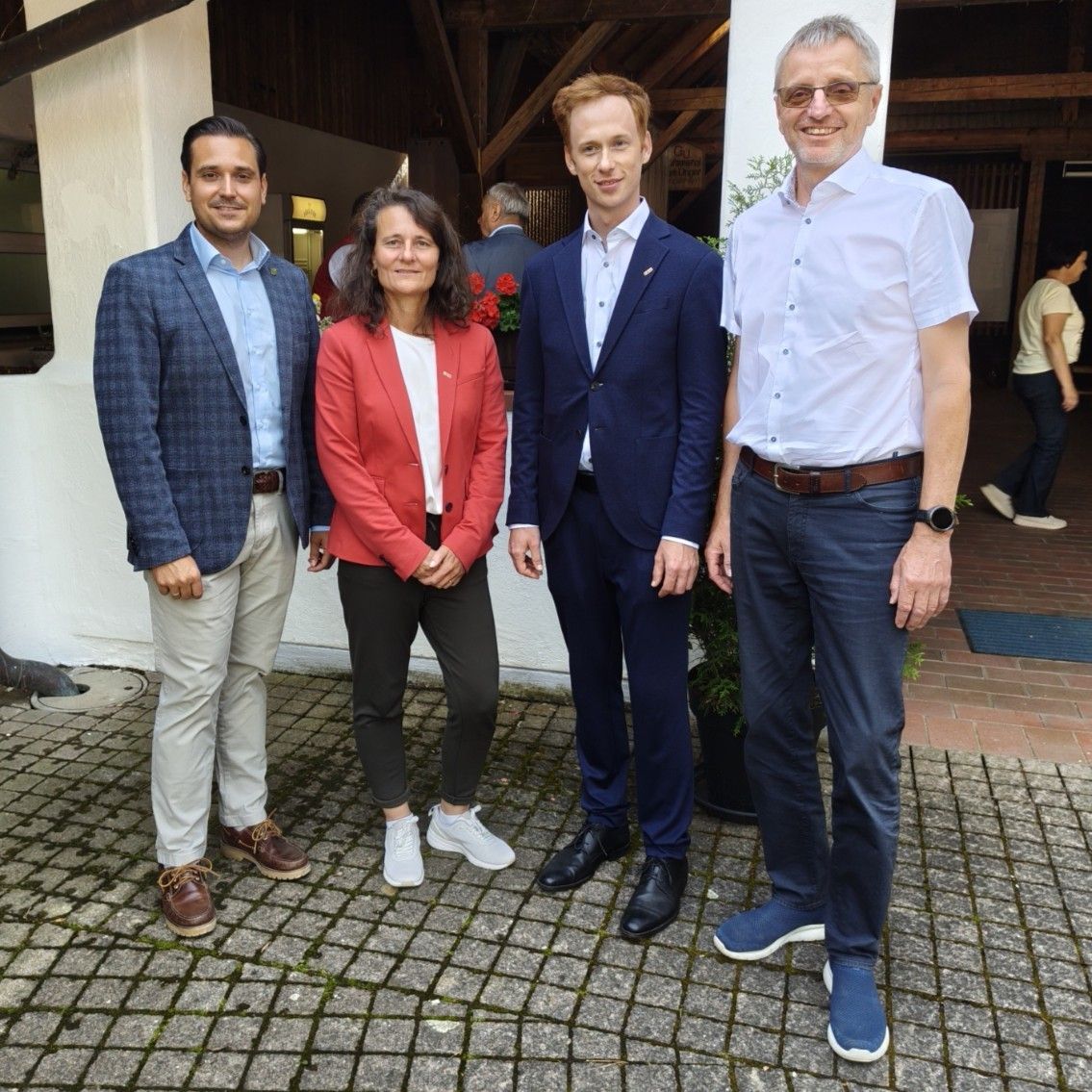 Four people are standing in front of a building. A woman in a red blazer and a man in a blue suit are in the center. They are all smiling and posing for a photo.