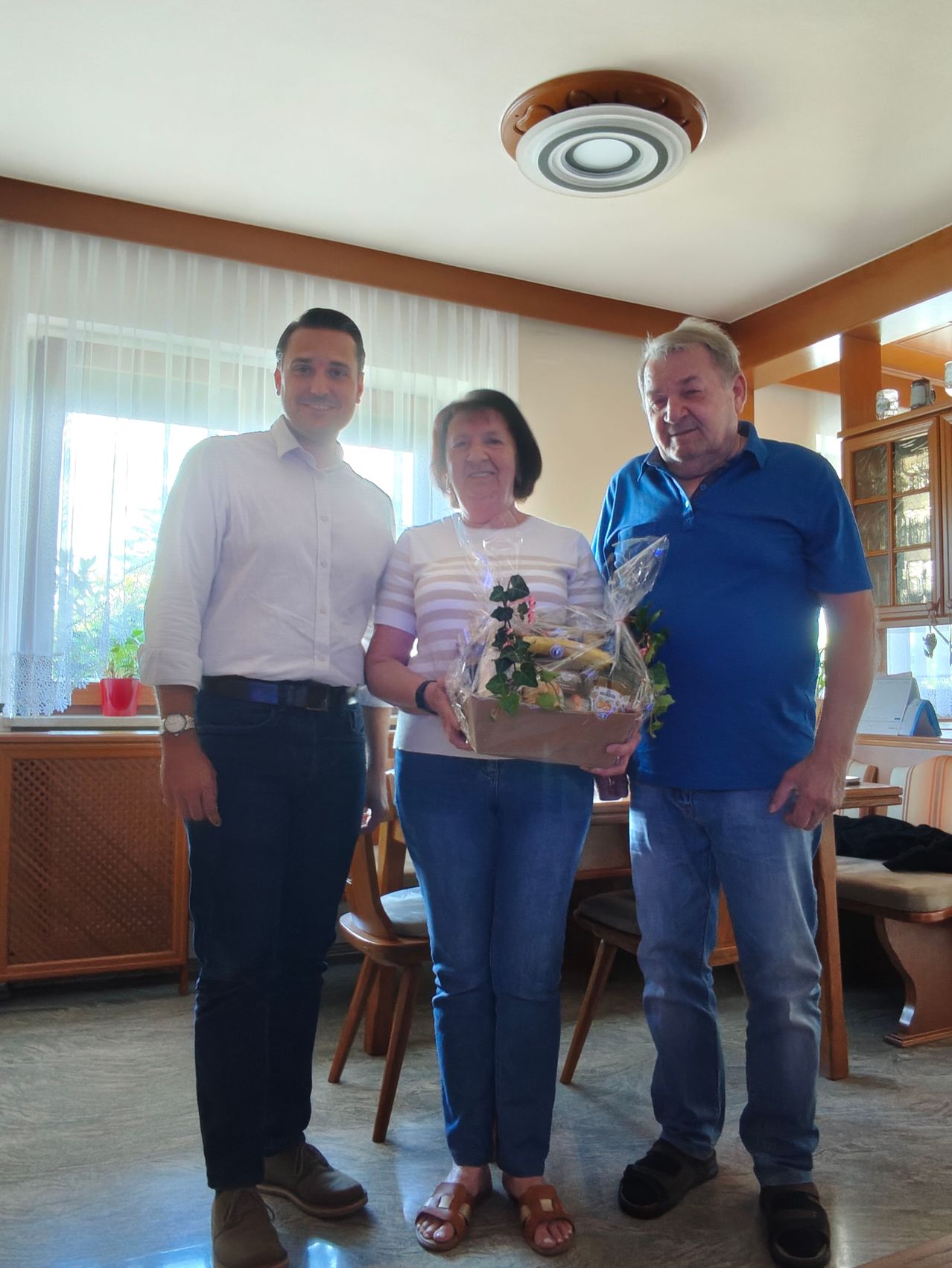 Three people stand in a room holding a flower arrangement. A man in a white shirt and a woman in a striped shirt hold a bouquet of flowers. Another man in a blue shirt stands beside them. Behind them, there are chairs, a table, and a cabinet.