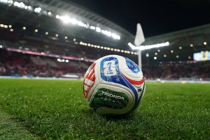 Ein Fußball mit dem FIFA-Logo liegt auf einem gut gepflegten Feld, mit einer Flagge in der Nähe und einem leeren Stadion im Hintergrund.