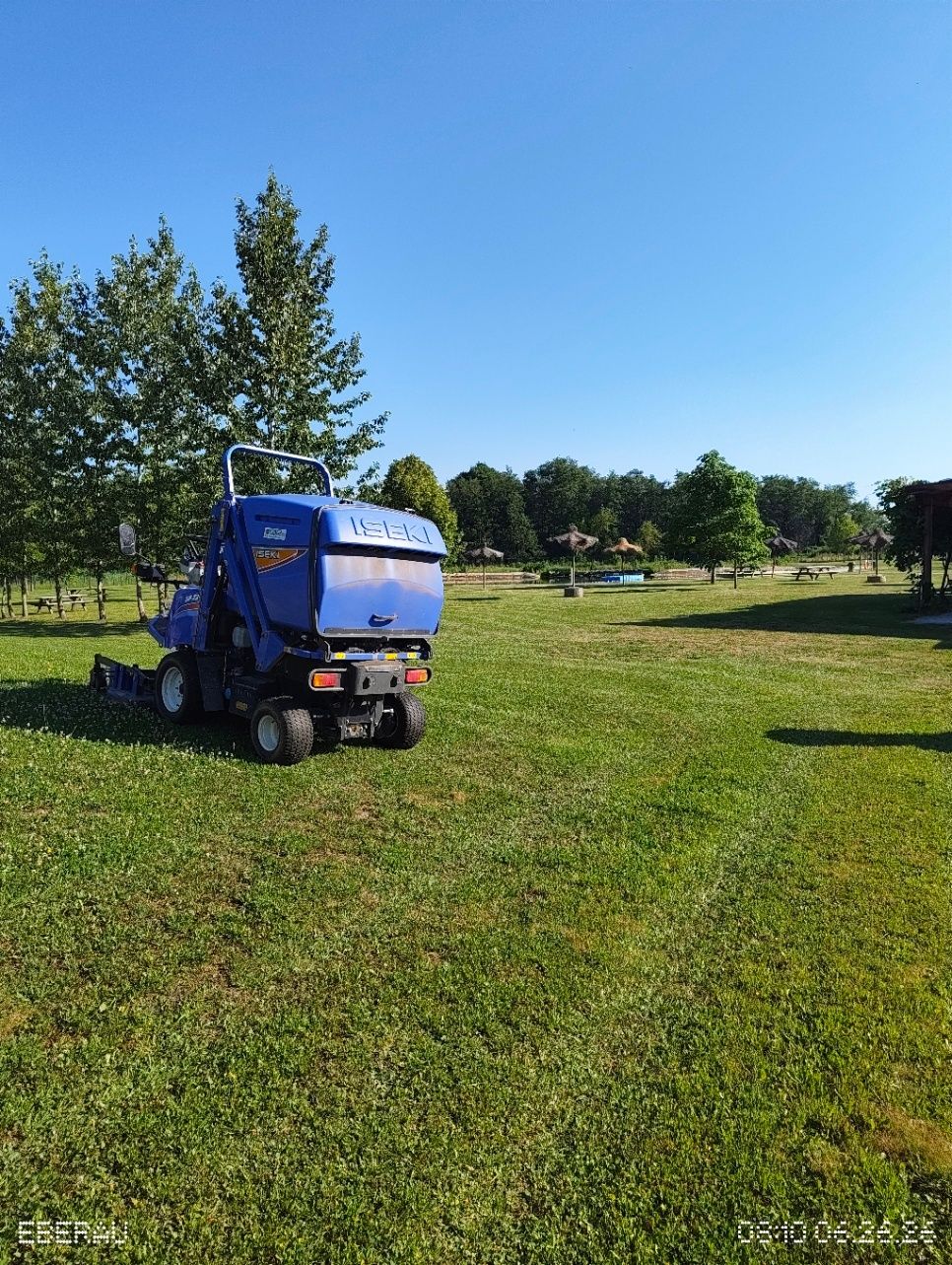 Ein blauer Rasenmäher steht auf einer grünen Wiese mit Bäumen und Picknickbereichen im Hintergrund an einem sonnigen Tag.