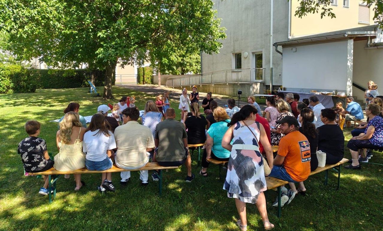 A group of people sit on benches in a grassy area, some standing, others listening, near a building with windows and a tree.