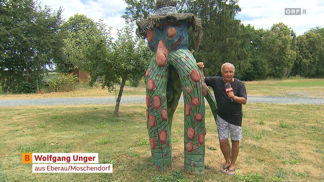A man in a black shirt and shorts stands next to a large, colorful wooden sculpture of a creature with a hat and tentacles in a grassy field.