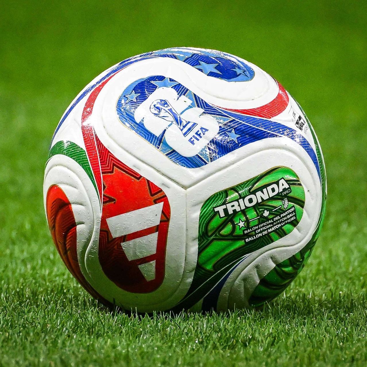 A soccer ball with a colorful design, featuring the FIFA logo, a red maple leaf, and the word 'TRIONDA' on a green background. It is placed on a green field.