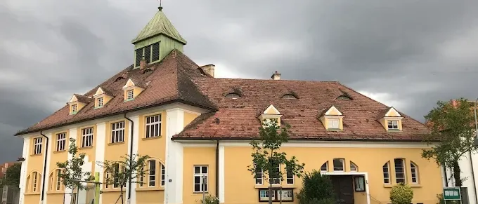 Ein gelbes Gebäude mit einem braunen Dach und einem Kreuz darauf, mit mehreren Fenstern und umgeben von Grün.