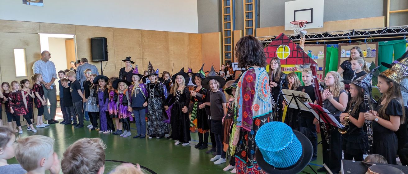 In einem Raum führen Kinder und Erwachsene in Kostümen eine musikalische Show auf. Eine Frau in farbenfroher Kleidung steht vor ihnen. Ein Basketballkorb ist an der Wand montiert.