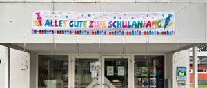 Ein farbenfrohes Banner mit den Worten 'Alles Gute zum Schulanfang' hängt über dem Eingang eines Gebäudes. Der Eingang hat zwei Glastüren und einen kleinen Gehweg. Es hängen Dekorationen über dem Eingang.