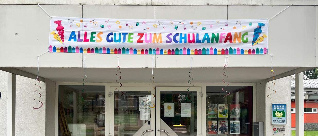 Ein farbenfrohes Banner mit den Worten 'Alles Gute zum Schulanfang' hängt über dem Eingang eines Gebäudes. Der Eingang hat zwei Glastüren und einen kleinen Gehweg. Es hängen Dekorationen über dem Eingang.