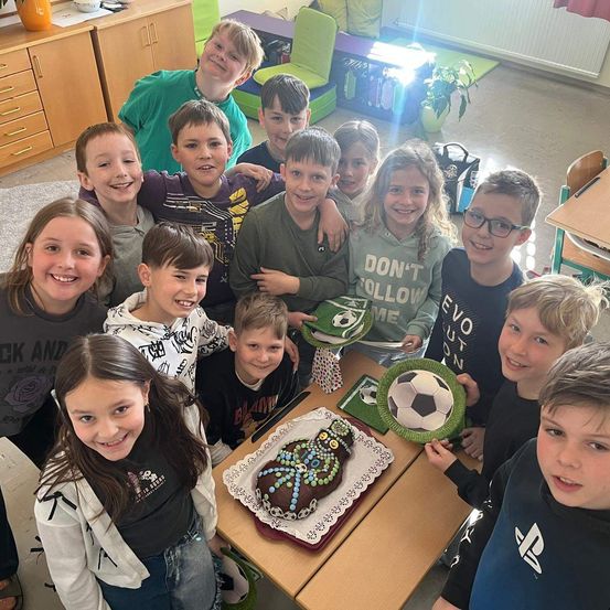 Eine Gruppe von Kindern in einem Klassenzimmer versammelt sich um einen Tisch mit einem Geburtstagskuchen. Einige von ihnen halten einen Fußball und tragen eine Brille. An der Seite stehen Schränke und eine Pflanze. Eine Person steht hinter ihnen und lächelt.