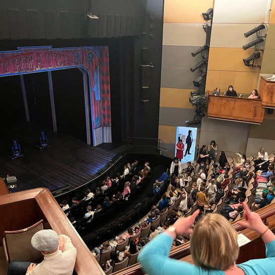 Ein Auditorium gefüllt mit einer Menschenmenge, einige tragen Hüte und andere haben Kinder dabei. Die Bühne hat ein farbenfrohes Design und eine Rampe.