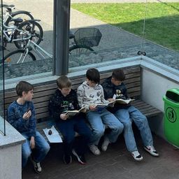 Vier Jungen sitzen auf einer Bank an einer Bushaltestelle. Sie tragen alle Freizeitkleidung und scheinen ein Buch zu lesen. Der Junge auf der linken Seite isst etwas. Hinter ihnen ist eine Glaswand mit Blick auf Fahrräder und Gras.