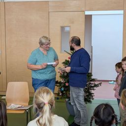 Eine Frau und ein Mann stehen vor einem Weihnachtsbaum, tauschen Papiere aus, umgeben von Kindern in einem Raum mit Holzwänden und einer Tür.