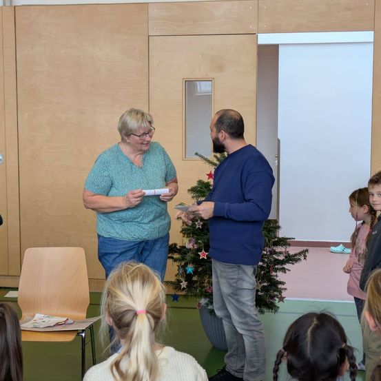 Eine Frau und ein Mann stehen vor einem Weihnachtsbaum, tauschen Papiere aus, umgeben von Kindern in einem Raum mit Holzwänden und einer Tür.
