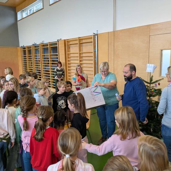 Eine Gruppe von Kindern und Erwachsenen versammelt sich in einem Raum. Eine Frau hält ein Papier mit bunten Buchstaben und ein Mann eine Flasche. Sie stehen in der Nähe eines Weihnachtsbaums.