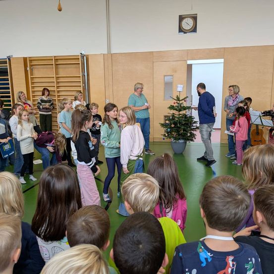 In einem Klassenzimmer versammeln sich eine Gruppe von Kindern und Erwachsenen um einen Weihnachtsbaum. Ein Mann steht in der Nähe und einige halten Instrumente. Eine Uhr ist an der Wand montiert.