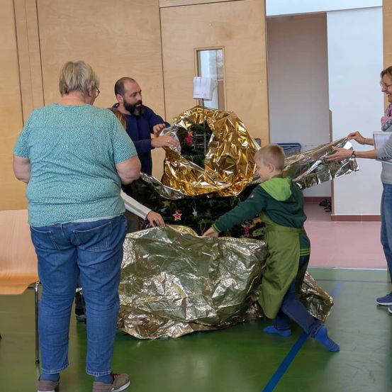 Mehrere Personen versammeln sich um einen geschmückten Weihnachtsbaum, der in Folie eingewickelt ist, in einem Raum. Eine Frau mit Brille hält ein eingewickeltes Geschenk. Ein Junge in einem grünen Mantel hilft, den Baum auszupacken.