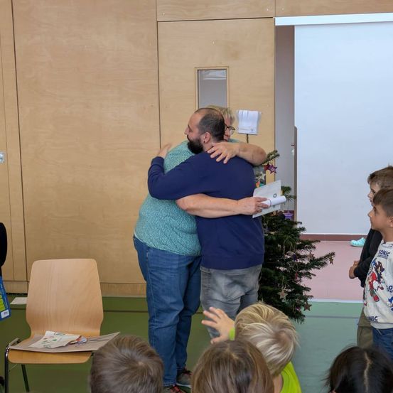 Zwei Erwachsene umarmen sich in einem Raum, in dem Kinder zusehen. Der Erwachsene im blauen Shirt hält ein Stück Papier. Hinter ihnen steht ein Weihnachtsbaum und eine Tür. Im Vordergrund befindet sich ein Stuhl mit Papieren.