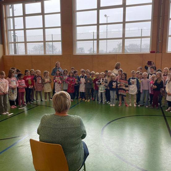 Eine Gruppe von Kindern in Schürzen steht in einer Reihe in einer Turnhalle, eine Frau sitzt vor ihnen auf einem Stuhl.