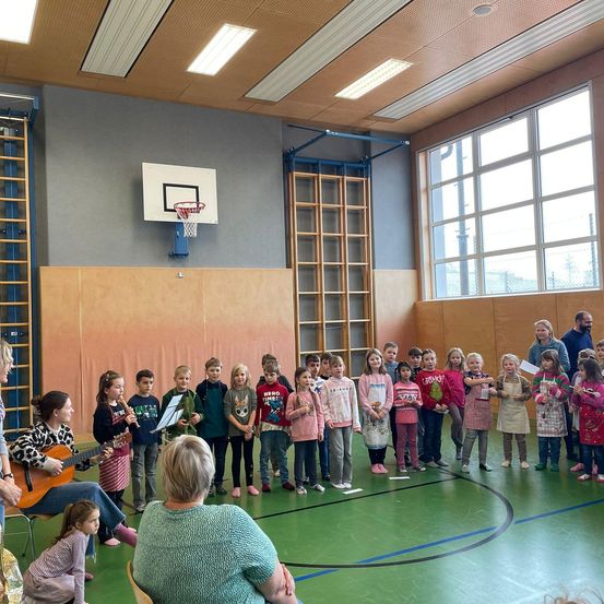 Eine Gruppe von Kindern und Erwachsenen befindet sich in einer Turnhalle, wobei einige Instrumente spielen und andere stehen. Eine Frau sitzt und spielt Gitarre. Ein Basketballkorb und große Fenster sind sichtbar.
