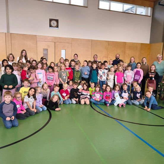 Ein Gruppenfoto von Kindern und Erwachsenen in einer Turnhalle mit einer Uhr an der Wand. Die Kinder tragen verschiedene Winterkleidung und einige tragen Schürzen. Sie sitzen und stehen auf dem grünen Boden.