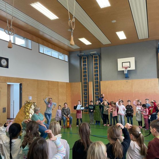 In einer Turnhalle nehmen Kinder und Erwachsene an einer Aktivität teil. Einige stehen, andere sitzen und einige halten Musikinstrumente. Eine Basketballkorb ist an der Wand montiert.