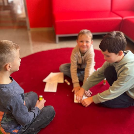 Drei Jungen sitzen auf einem roten Teppich und sind in einer Aktivität verwickelt. Einer hält ein Stück Papier, während ein anderer einen Marker hält. Sie lächeln und schauen sich gegenseitig an.