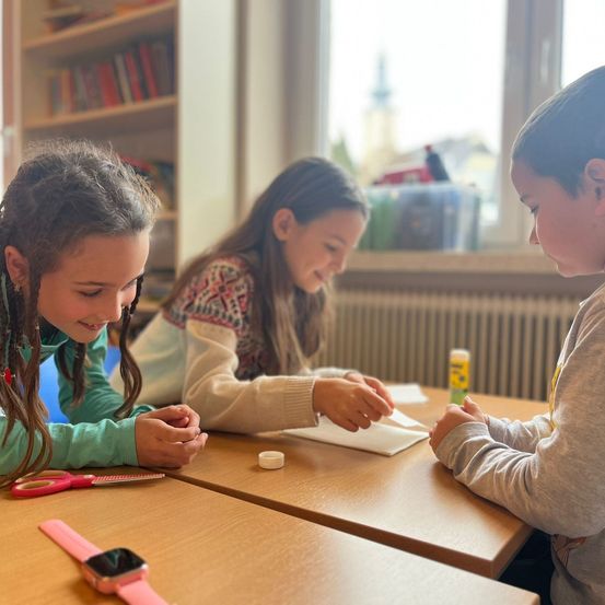Drei Kinder arbeiten in einem Klassenzimmer an einem Bastelprojekt an ihren Schreibtischen mit Blick auf einen Kirchturm außerhalb des Fensters.