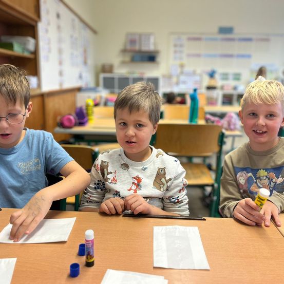 Drei Jungen sitzen in einem Klassenzimmer an einem Tisch, einer hält einen Klebestift und Papier. Ein anderer Junge hat eine Zeichnung auf dem Arm. Verschiedene Gegenstände wie Klebstoffflaschen und Papier liegen auf dem Tisch.