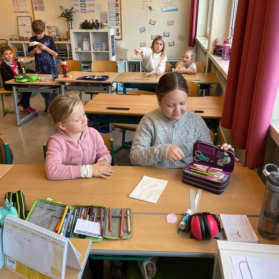 In einem Klassenzimmer sitzen Kinder an Tischen, eine Mädchen öffnet einen Stiftetui. Eine andere Mädchen hält ein Papier. Verschiedene Gegenstände sind auf den Tischen, darunter Notizbücher, Stifte und ein rotes Objekt.