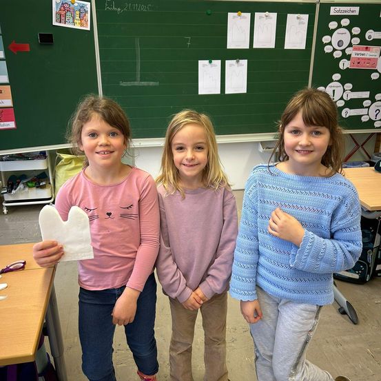 Drei junge Mädchen stehen lächelnd in einem Klassenzimmer nebeneinander und posieren für die Kamera. Jedes von ihnen hält ein Stück Papier, wobei eine ein weißes katzenförmiges Objekt hält. Hinter ihnen steht eine Tafel mit verschiedenen Zeichnungen und Texten.