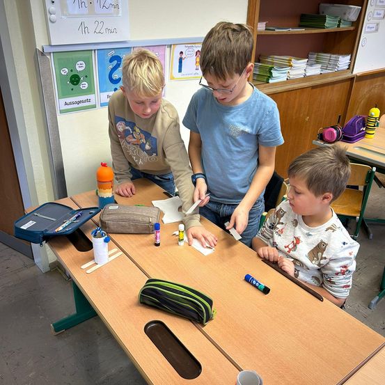 Drei Jungen sind in einer Aktivität an einem Schreibtisch beschäftigt, umgeben von Schulmaterialien. Ein Junge hält ein Papier und eine Schere, während ein anderer einen Marker benutzt. Verschiedene Gegenstände wie Stifte, Marker und ein Bleistiftetui stehen auf dem Schreibtisch.