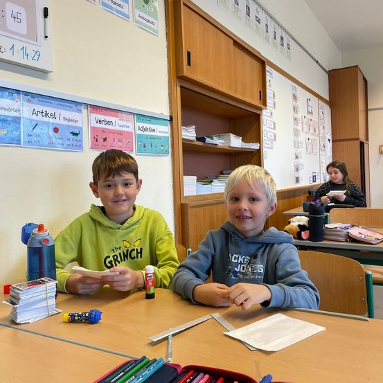 Zwei Jungen sitzen an einem Schreibtisch in einem Klassenzimmer mit verschiedenen Gegenständen auf dem Schreibtisch. Eine Lehrerin sitzt am anderen Schreibtisch im Hintergrund.