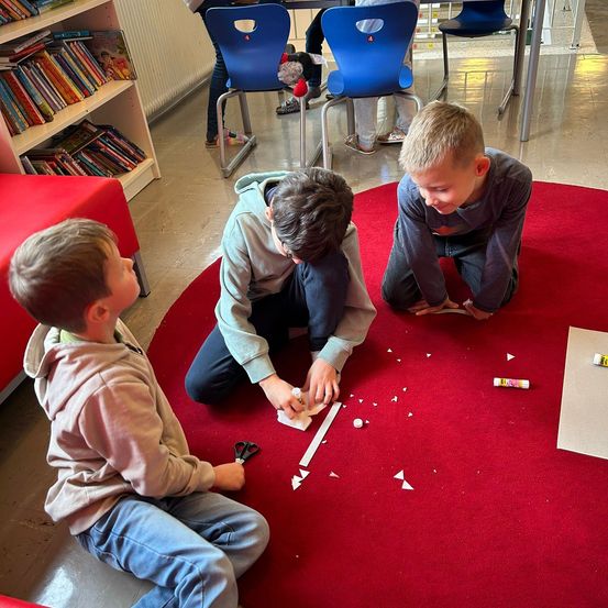 Drei Jungen sitzen auf einem roten Teppich und sind in ein Projekt vertieft. Einer hält Scheren, ein anderer eine Flasche und der dritte beobachtet. Dahinter steht ein Bücherregal und Stühle.