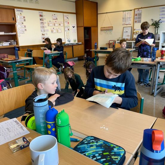 In einem Klassenzimmer sitzen Kinder an Schreibtischen. Einige lesen, andere schreiben. Flaschen, ein Becher und ein Rucksack liegen auf dem Schreibtisch. Ein Junge steht am Tisch. Papiere hängen an der Wand.