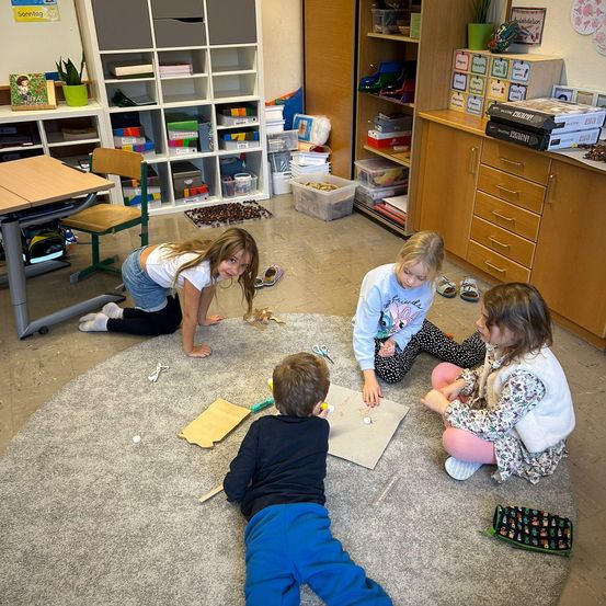 Vier Kinder spielen auf einem Teppich in einem Klassenzimmer. Sie basteln mit Papier und Scheren. Regale und Schränke im Hintergrund.