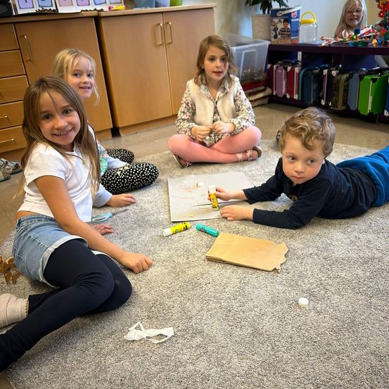 Vier Kinder sitzen auf dem Boden in einem Klassenzimmer und beschäftigen sich mit einem Kunstprojekt. Ein Junge liegt auf dem Bauch und hält ein Stück Papier.