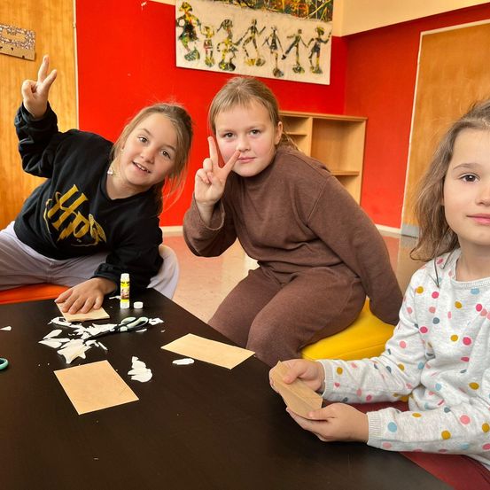 Drei junge Mädchen sitzen um einen Tisch herum und basteln. Zwei der Mädchen geben Friedenszeichen. Auf dem Tisch liegen Papiere und Scheren.