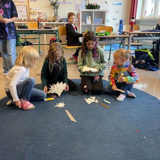 Fünf Kinder sitzen auf dem Boden und beschäftigen sich mit einer Bastelaktivität. Vier Mädchen arbeiten an Papierbasteleien, während ein Junge auf einem Stuhl beobachtet. Der Raum hat Tische, Stühle und eine Topfpflanze.
