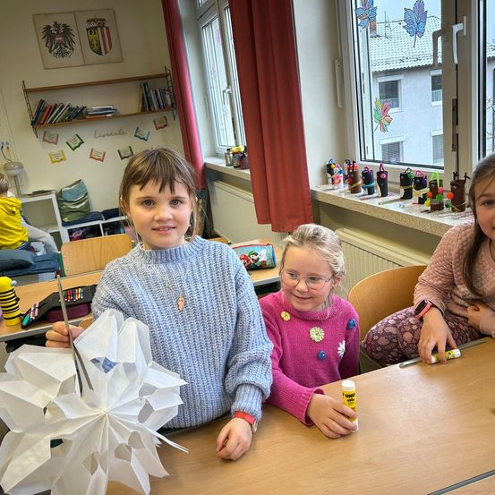 Drei Mädchen sitzen an einem Schreibtisch und beschäftigen sich mit einer Bastelaktivität. Das Mädchen in der Mitte hält eine große Papierschneeflocke. Das Mädchen auf der rechten Seite trägt eine Brille und hält einen Klebestift. Hinter ihnen befindet sich ein Fenster mit Dekorationen auf der Fensterbank.