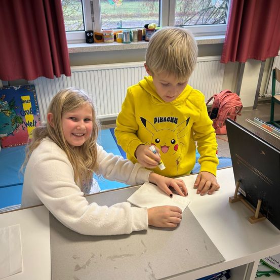 Zwei Kinder sitzen an einem Schreibtisch in einem Klassenzimmer. Das Mädchen benutzt einen Marker, um auf ein Stück Papier zu schreiben. Der Junge hält einen Marker. Hinter ihnen befindet sich ein Fenster mit Blick auf das Freie.