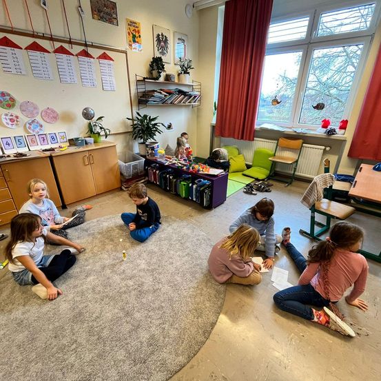 In einem Klassenzimmer sitzen Kinder auf dem Boden um einen Kreis herum und beschäftigen sich mit Aktivitäten. Topfpflanzen und Bücher schmücken den Raum. Ein großes Fenster bietet einen Blick auf Bäume draußen.