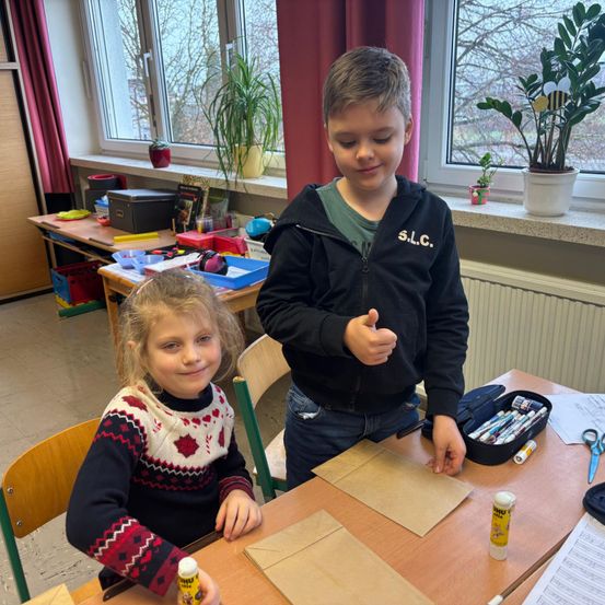 Zwei Kinder in einem Klassenzimmer, ein Mädchen mit einem gemusterten Pullover und ein Junge in einer schwarzen Kapuzenjacke, stehen bei einem Tisch mit Bastelutensilien, lächeln und schauen sich etwas auf dem Tisch an.