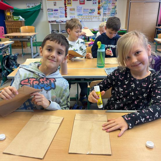 In einem Klassenzimmer sitzen Kinder an Tischen, wobei sich zwei Jungen und ein Mädchen auf das Basteln konzentrieren. Der Junge auf der linken Seite hält einen braunen Papierbeutel, während das Mädchen auf der rechten Seite einen Klebestift in der Hand hat. Verschiedene Gegenstände wie eine Flasche und Papiere befinden sich auf den Tischen hinter ihnen.