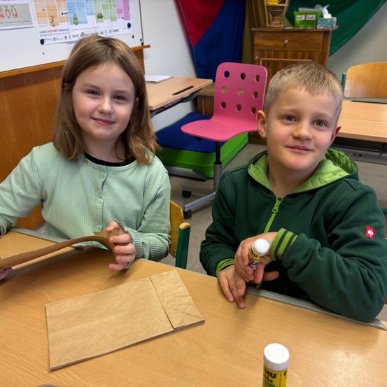 Zwei Kinder, ein Junge und ein Mädchen, sitzen an einem Tisch in einem Klassenzimmer. Sie lächeln und scheinen an einem Bastelprojekt zu arbeiten. Das Mädchen hält einen braunen Papierbeutel und einen Stock, während der Junge eine Flasche Kleber in der Hand hat. Der Raum ist mit bunten Stühlen, Tischen und einer Pinnwand mit verschiedenen Postern ausgestattet.