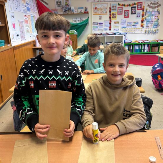 Zwei Jungen in einem Klassenzimmer, einer hält einen braunen Papierbeutel und der andere einen Klebestift. Hinter ihnen sitzen andere Kinder an Schreibtischen. Das Klassenzimmer ist mit verschiedenen Gegenständen dekoriert.
