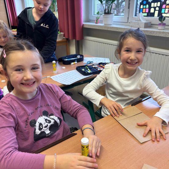 Zwei Mädchen sitzen lächelnd an einem Schreibtisch im Klassenzimmer. Eine trägt ein lila T-Shirt mit Panda-Grafik, die andere in Weiß. Sie haben Bastelmaterial vor sich auf dem Schreibtisch.