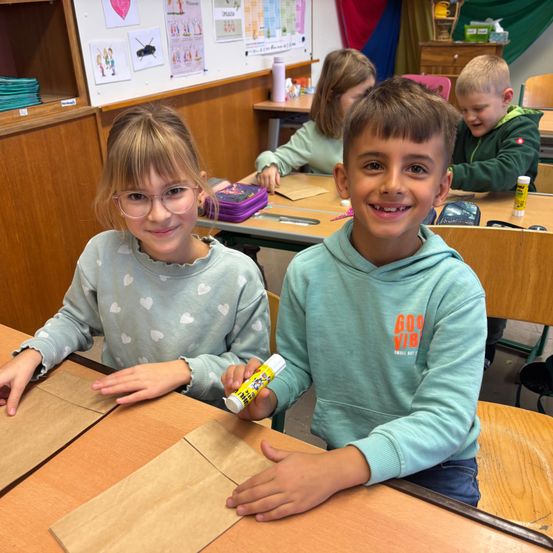 Zwei Kinder lächeln in die Kamera, während sie in einem Klassenzimmer ein Bastelprojekt bearbeiten. Das Mädchen trägt eine Brille und der Junge hält einen Klebestift.