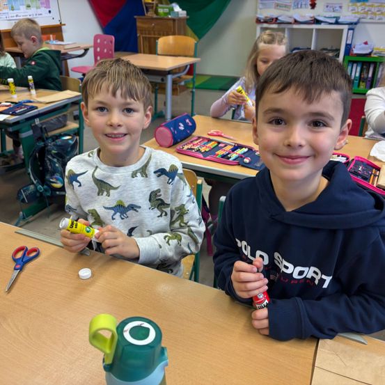 Zwei Jungen sitzen an einem Schreibtisch im Klassenzimmer, einer hält einen Klebestift und lächelt. Der andere hält eine Klebeflasche und lächelt. Auf dem Tisch liegen eine Schere, ein Stiftetui und eine Wasserflasche.