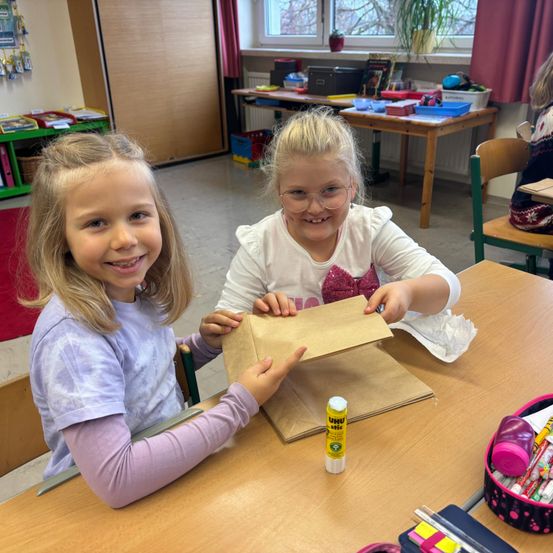 Zwei junge Mädchen sitzen in einem Klassenzimmer an einem Schreibtisch, halten einen braunen Papierbeutel und lächeln. Das Mädchen links trägt ein lila Langarmshirt. Ein Klebestift und andere Klassenzimmergegenstände sind auf dem Schreibtisch.