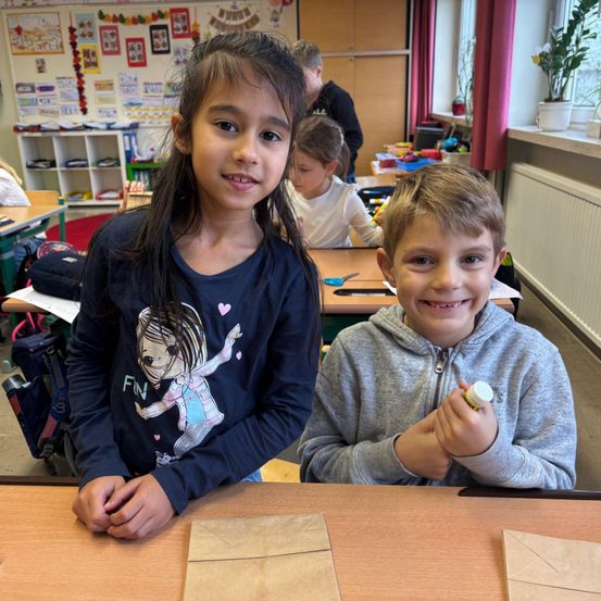 Zwei Kinder, ein Mädchen und ein Junge, lächeln und sitzen an einem Tisch im Klassenzimmer. Das Mädchen hat ein blaues Shirt mit einem Cartoon-Charakter, und der Junge trägt eine graue Kapuzenjacke. Hinter ihnen sitzen andere Kinder an ihren Tischen.