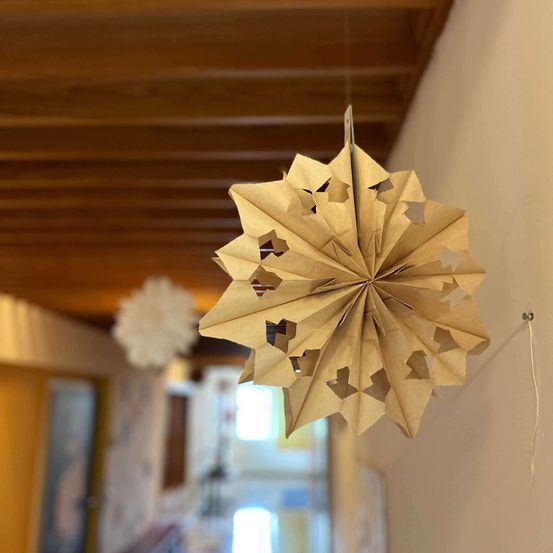 Eine braune Papier-Schneeflocke hängt von der Decke, möglicherweise in einem Flur oder Eingangsbereich. Links hängt eine weiße Papier-Schneeflocke.